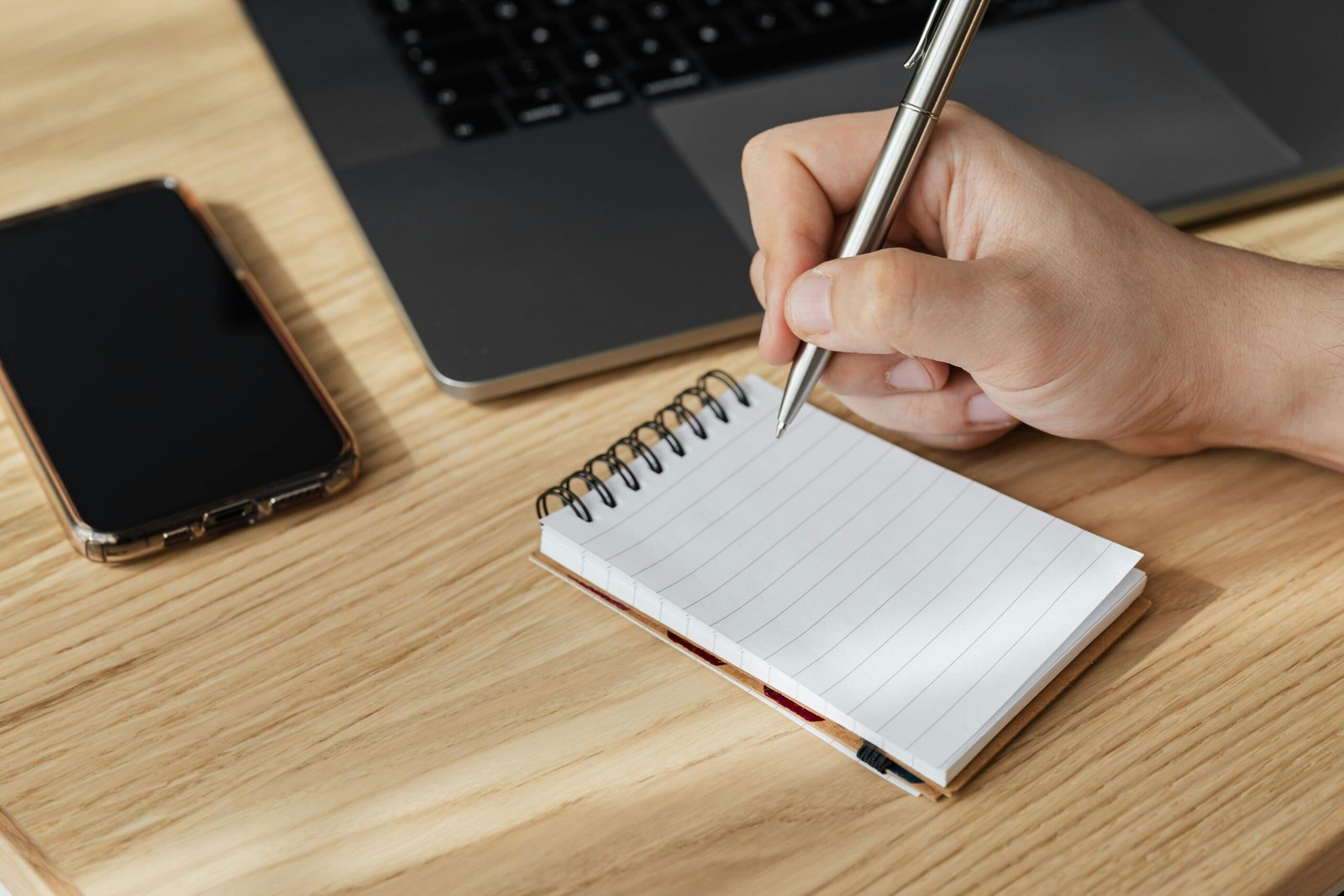 A person writing in a notepad next to a laptop and smartphone on a wooden desk.
